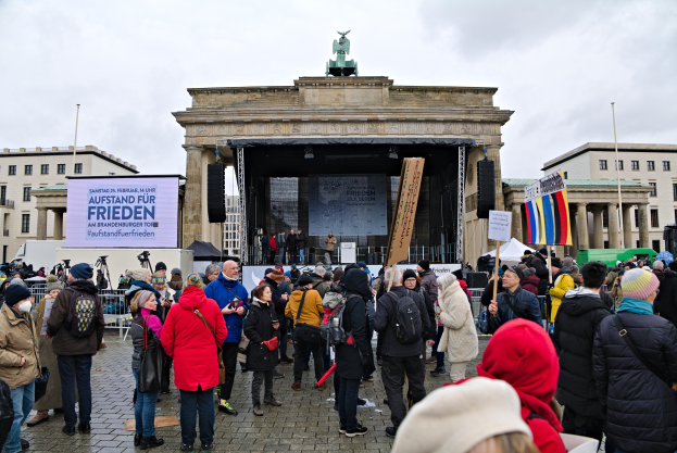 Eine Menschenmenge steht vor einem Gebäude mit einer Bühne, auf der Redner und ein Bildschirm zu sehen sind, mit Flaggen und Transparenten auf der rechten Seite, was auf eine Demonstration in Berlin hindeutet.