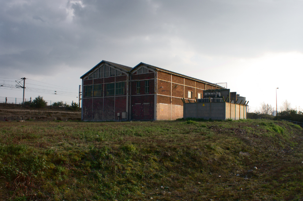 Eine Szene mit Schuppen, Transformatoren, Strommasten mit Kabeln, Zäunen, einem Straßenmast mit Laterne, Bäumen und einem bewölkten Himmel.