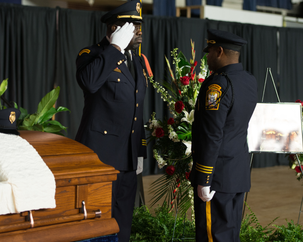 Zwei Polizisten in Uniform stehen neben einem Sarg, der mit einem weißen Tuch bedeckt ist.