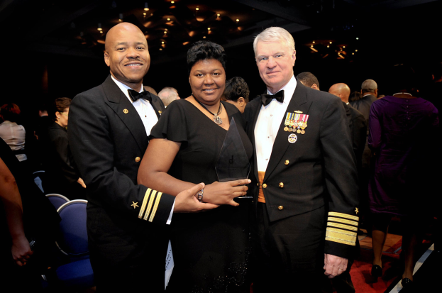 Zwei Männer in schwarzen Blazern und weißen Hemden stehen neben einer Frau in einem schwarzen Kleid, die eine Auszeichnung hält, auf einem roten Teppich, während andere sitzen und stehen in einer beleuchteten Innenraumkulisse.