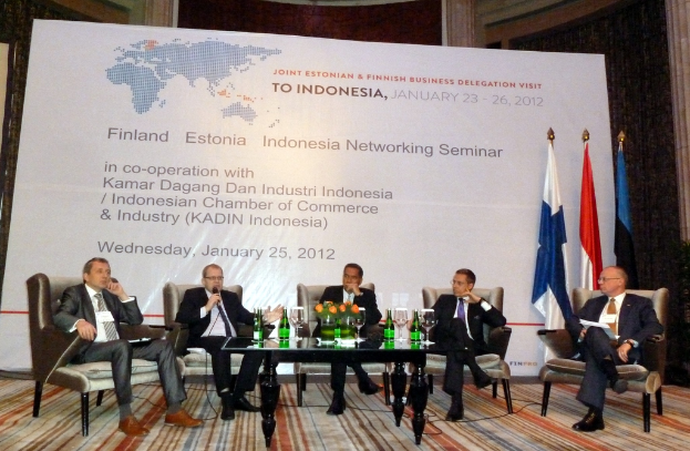 Eine Bühne mit Menschen, Stühlen, einem Mikrofon, Kabeln, einem Tisch, Flaschen, Blumen, Gläsern und anderen Gegenständen, mit Flaggen und einem Banner in der Mitte und einem Gebäude mit Vorhängen im Hintergrund.