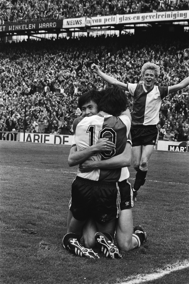 Drei Fussballspieler feiern ein Tor auf dem Boden, umgeben von einer jubelnden Menge und schwenkenden Fahnen im Hintergrund, dargestellt in Schwarz-Weiss.