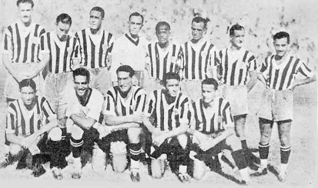 Ein Schwarz-Weiß-Foto einer Fussballmannschaft, mit einigen stehenden und anderen sitzenden Spielern, mit Text unten.