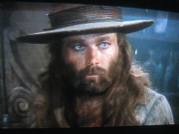 Ein Mann mit Bart und Schnurrbart trägt einen Hut und ist auf einem Bildschirm zu sehen.