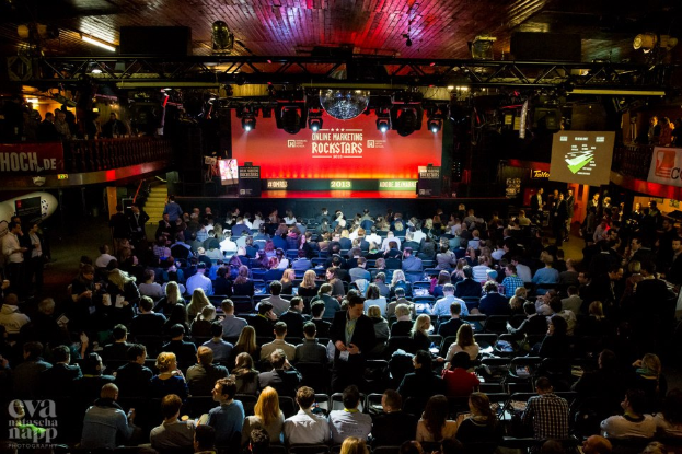 Eine große Menschenmenge sitzt auf Stühlen vor einer Bühne mit Bildschirmen und Bannern, die von Scheinwerfern beleuchtet wird und von Geländern umgeben ist, mit einem Wasserzeichen in der linken unteren Ecke, das eine Atmosphäre von Aufregung und Erwartung für ein Online-Marketing-Rockstars-Event schafft.
