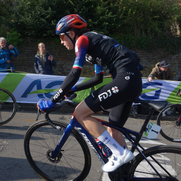 Eine professionelle Radrennfahrerin, Katie Archibald, trägt einen Helm, eine Brille, Handschuhe und Schuhe und fährt ein Fahrrad auf einer Straße an einer Menschenmenge vorbei, mit einem Banner, einer Wand, Bäumen und einem Gebäude im Hintergrund.