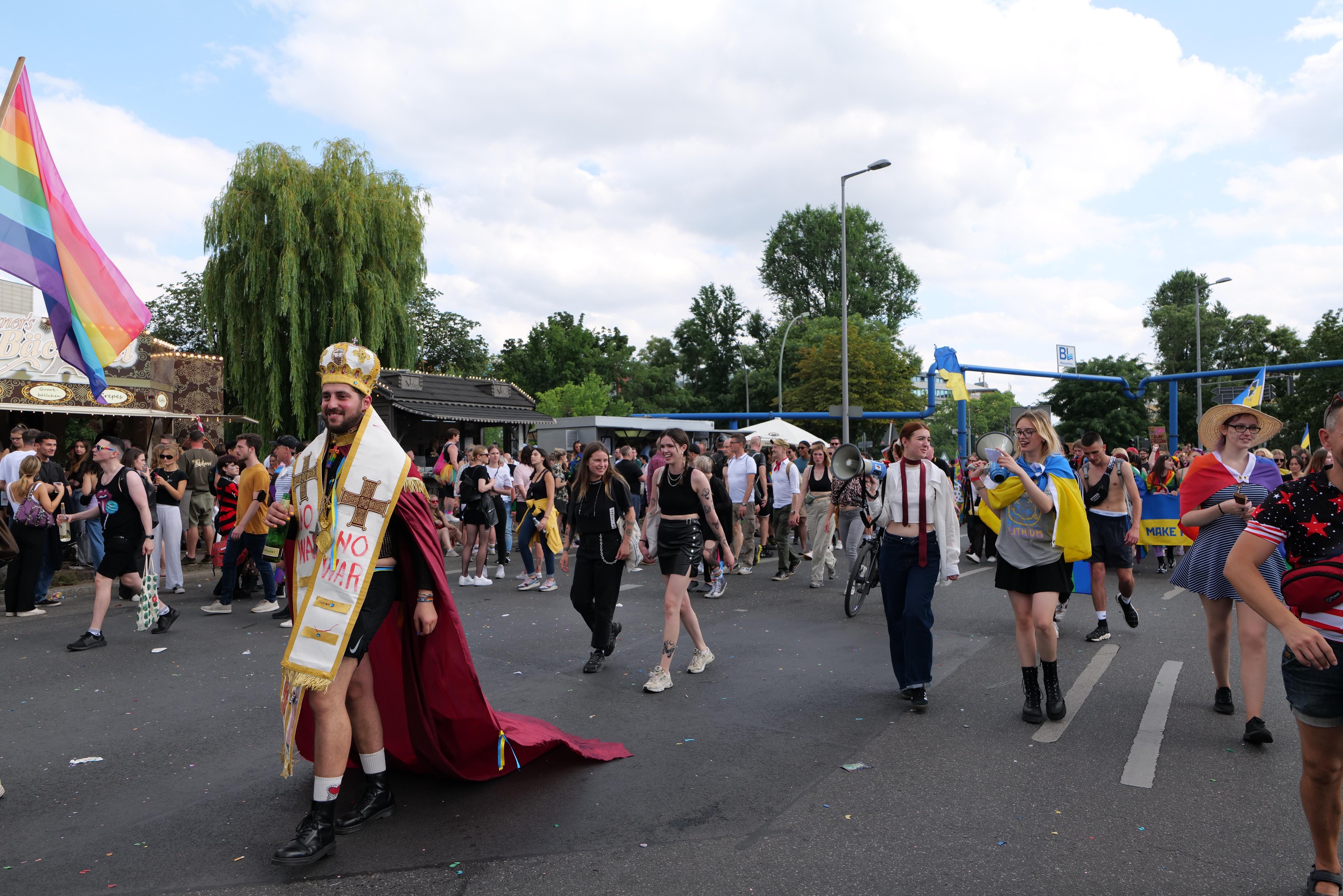 Eine Gruppe von Menschen marschiert bei der Gay Pride Parade 2018 mit einer Regenbogenflagge und Musikinstrumenten, einige tragen Mützen, vor einem Hintergrund aus Laternenmasten, Bäumen, Schuppen und einem bewölkten Himmel.