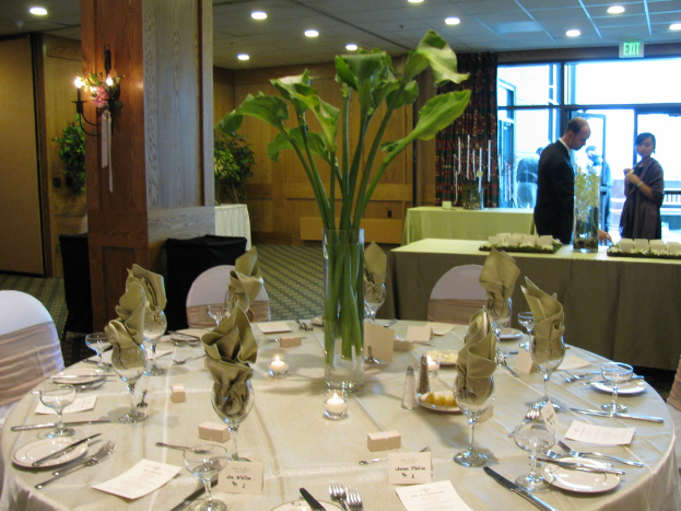 Ein weißes Tischtuch mit Tellern, Besteck und einem Glas, umgeben von Stühlen, mit einem grünen Tisch dahinter; ein Mann in einem schwarzen Blazer und eine Frau stehen in der Nähe einer Tür mit zwei Männern dahinter.