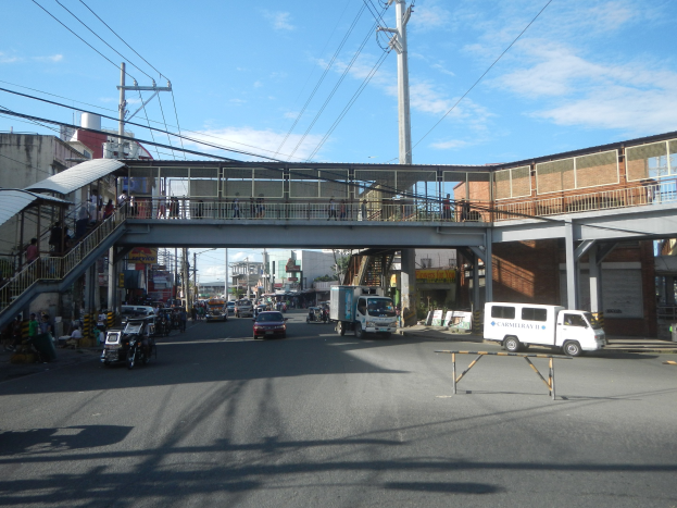 Stadtstraße mit Fahrzeugen, eine Fußgängerbrücke mit Menschen, Strommasten, Gebäuden und einem bewölkten Himmel.