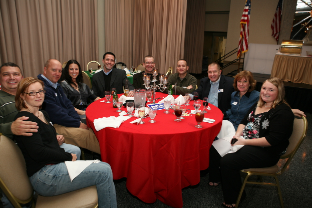 Eine Gruppe strahlender Menschen an einem Tisch mit Gläsern, Papieren, Weinflaschen und Flagge auf der rechten Seite und Vorhängen an der Wand.