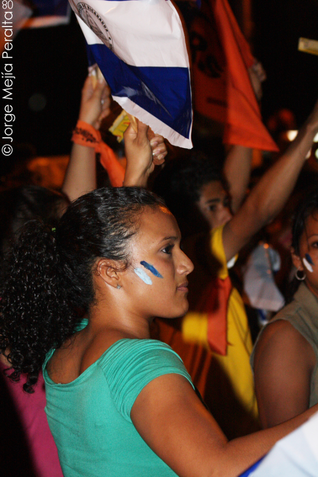 Eine Frau in einem grünen T-Shirt steht auf einer Demo und hält ein weißes Schild hoch, während viele andere Menschen hinter ihr stehen.