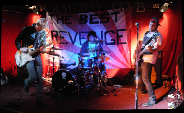 Zwei Personen vor einem Mikrofon mit Gitarren stehend, eine Person sitzt und spielt Schlagzeug mit Brille, vor einem Hintergrund mit hängenden Flexi-Schallplatten und CDs.