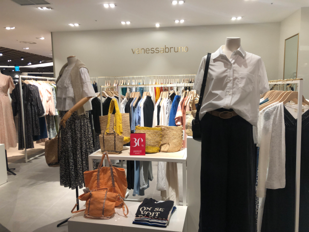 Ein Vanessa Bruno Geschäft in Singapur mit Schaufensterpuppen in Kleidern, Kleidung an Bügeln, Taschen und anderen Gegenständen auf Tischen, einem Textschild an der Wand und einer Deckenbeleuchtung.