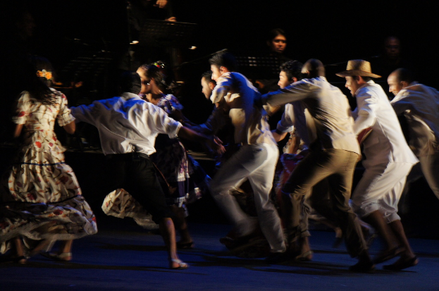 Eine Gruppe von Menschen tanzt im Vordergrund mit einer anderen Gruppe von Menschen, Ständen und Mikrofonen im Hintergrund, alles auf einem Boden.