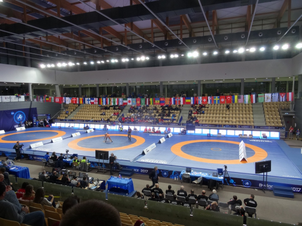 Großer Innenstadionsaal mit Zuschauern bei einem Wrestling-Match, einschließlich sitzender und stehender Zuschauer, Tischen mit Gegenständen und Hintergrundflaggen, Lichtern und Stangen.