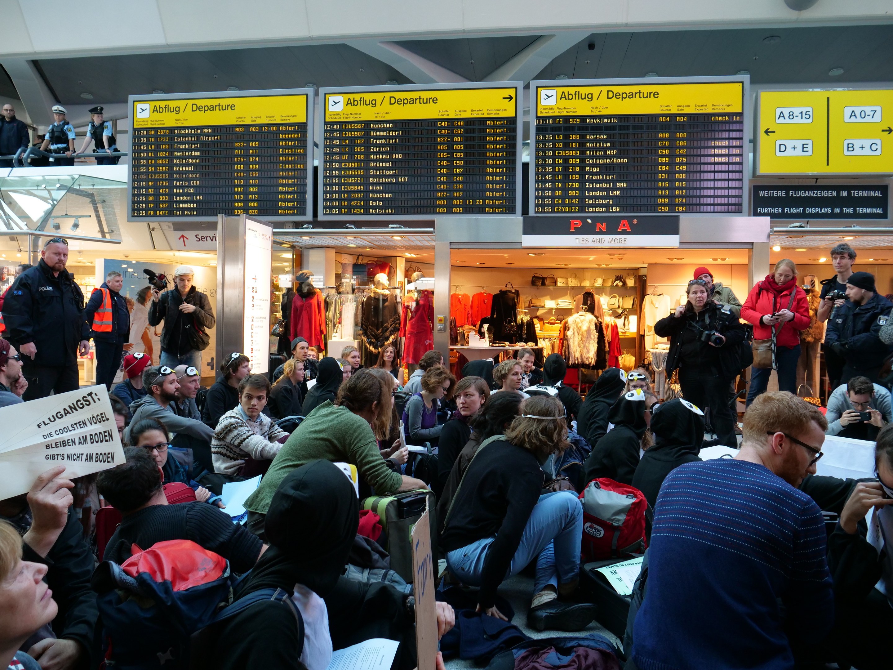 Große Gruppe von Menschen sitzt und steht in einem Flughafen während einer Demonstration, mit Informationsschildern, Schaufensterpuppen in Kleidern und Deckenbeleuchtung im Hintergrund.