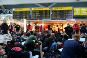 Große Gruppe von Menschen sitzt und steht in einem Flughafen während einer Demonstration, mit Informationsschildern, Schaufensterpuppen in Kleidern und Deckenbeleuchtung im Hintergrund.