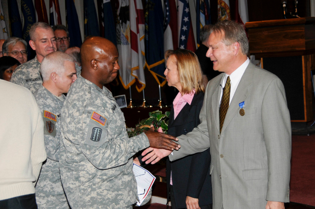 Eine Gruppe von Menschen mit zwei Personen, die Hände schütteln, im Hintergrund Fahnen an Stangen, ein Podium mit Mikrofon und eine Topfpflanze in der Nähe.