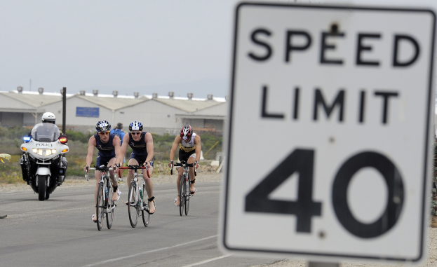 Gruppe von Radfahrern mit Helmen auf einer Straße mit einem Geschwindigkeitsbegrenzungsschild, Gebäuden und Bäumen im Hintergrund unter einem klaren blauen Himmel.