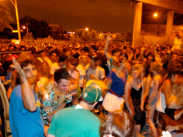 Leute tanzen im Vordergrund eines Konzerts, mit Lichtern, Masten, Bäumen und anderen Gegenständen im Hintergrund.