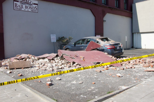 Eine eingestürzte Wand mit verstreuten roten Ziegeln, ein geparktes Auto, eine Topfpflanze und ein Gebäude mit einer Tafel, auf der 'Kautionsfreiheit' steht.