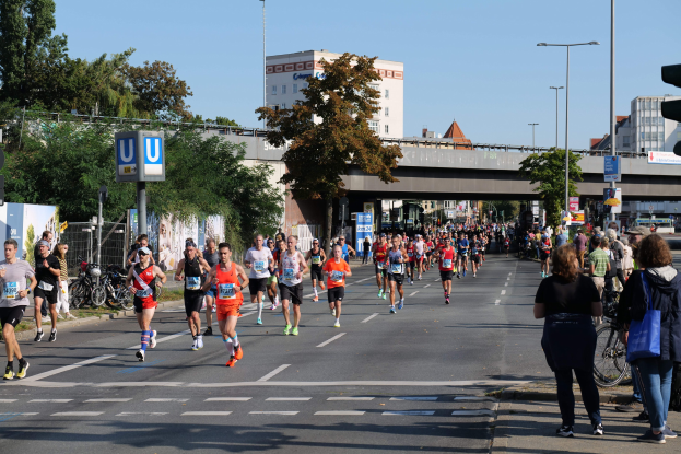 Gruppe von Menschen beim Laufen auf einer von Bäumen gesäumten Straße mit Laternen, Schildern, Fahrrädern, einem Zaun, Gras, einer Brücke, Gebäuden und einem klaren blauen Himmel.