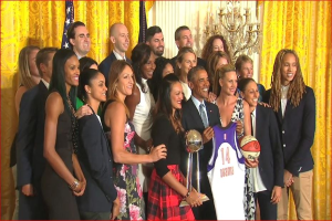 Präsident Obama und First Lady Michelle Obama posieren mit dem Damen-Basketball-Team im Oval Office des Weißen Hauses, halten einen Basketball, eine Trophäe und lächeln in der Nähe einer Flagge, Vorhänge und eines Kerzenleuchters.
