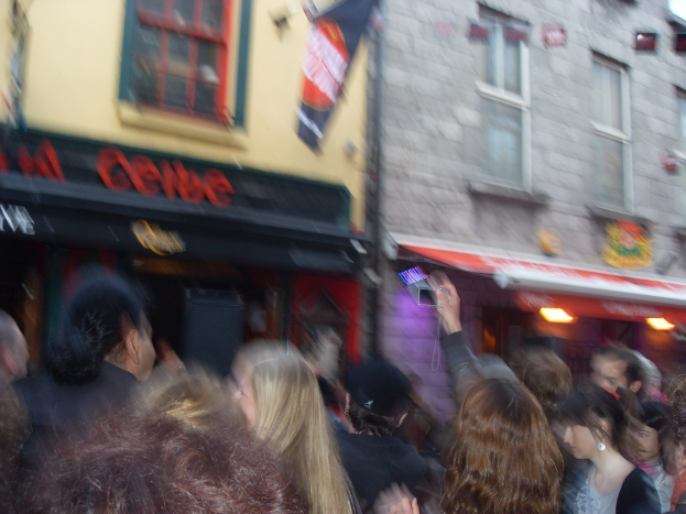 Ein unscharfer Bildausschnitt, der unten Menschen auf der StraÃŸe und oben GebÃ¤ude mit Lichtern, Fenstern und einer Fahne zeigt.