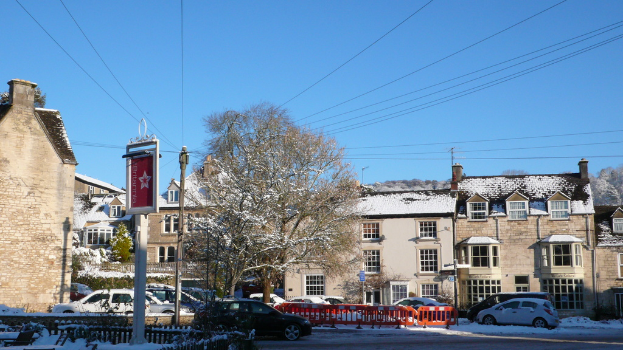 Fahrzeuge auf einer schneebedeckten Straße mit Häusern, Bäumen und Strommasten im Hintergrund, alles unter Schnee begraben.
