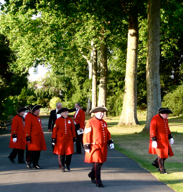 Eine Gruppe älterer Männer in einheitlicher Kleidung, wobei ein Mann in einem Hemd, Blazer, Hose und Schuhen auf einer Straße steht, mit Gebäuden, Bäumen, Pflanzen und Gras im Hintergrund.