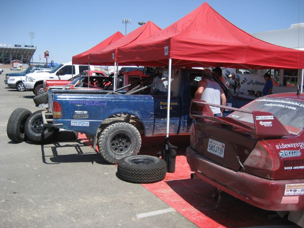 Mehrere Fahrzeuge mit sichtbaren Reifen sind auf einem roten Teppich geparkt, mit Menschen in der Nähe. Rote Zelte sind im Hintergrund aufgebaut, zusammen mit Straßenlaternen und einem Schuppen auf der linken Seite, unter einem klaren Himmel.