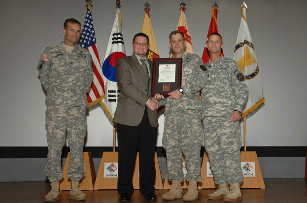 Vier Männer stehen auf einer Bühne: drei in Militäruniformen und einer in Blazer, Krawatte und Hemd, die Hände schütteln und einen Preis überreichen, mit sechs Fahnen auf Ständern und einer weißen Wand im Hintergrund.