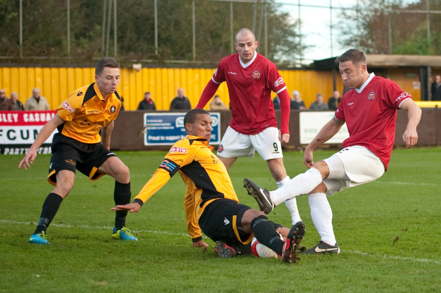 Spieler in blauen und roten Uniformen spielen ein Spiel auf einem grasbewachsenen Feld mit einem Ball, während Zuschauer außerhalb des Spielfelds stehen und sie anfeuern, mit einem Baum und Himmel im Hintergrund.