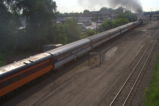 Ein Zug auf Schienen mit Bäumen, Strommasten mit Kabeln, Autos und Gebäuden links und eine Brücke vorne, die Rauch abgibt.