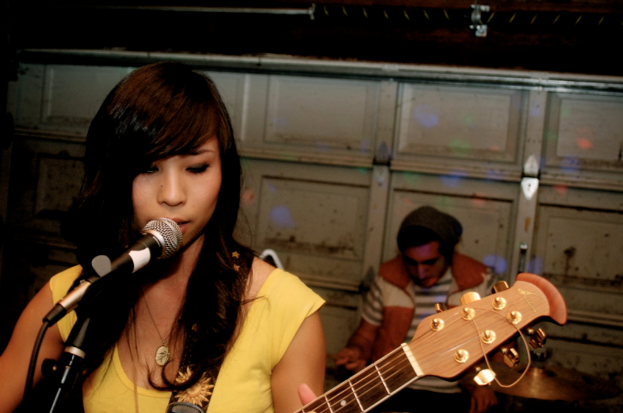 Eine Frau in der Mitte spielt Gitarre und singt in ein Mikrofon links, während ein sitzender Mann im Hintergrund rechts zu sehen ist, mit einer Wand dahinter.