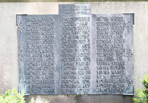 Ein Steinplaque an einer Wand mit eingravierter Schrift, umgeben von Pflanzen unten, wahrscheinlich ein Holocaust-Mahnmal.