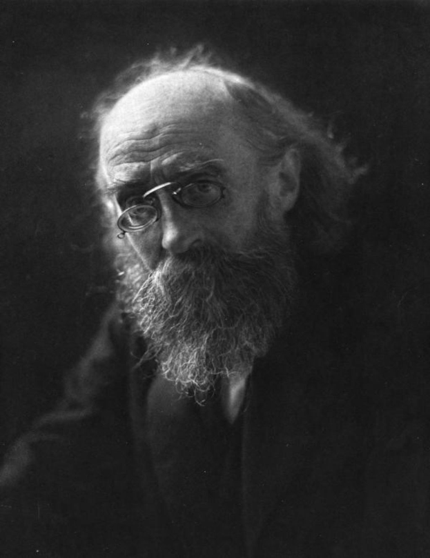 Schwarz-weiß-Porträt eines älteren Mannes mit langem Bart und Brille vor dunklem Hintergrund.