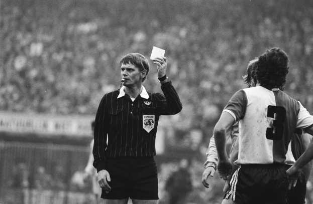 Ein Schiedsrichter in Schwarz-Weiß-Händler hält eine rote Karte vor einer Menge von Menschen in Füßballkleidung, mit einem Zaun im Hintergrund.