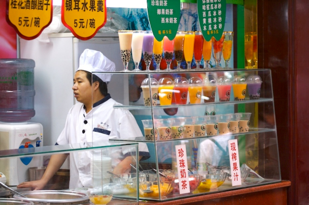 Ein Koch steht vor einem Tisch mit Saftgläsern, Schalen mit Süßigkeiten und Bannern, mit einem Trinkwassergerät und Kühlschrank im Hintergrund.
