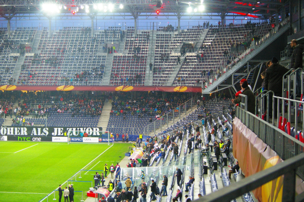 Großes Stadion mit Zuschauern bei einem Fußballspiel, einige stehen und sitzen auf Stühlen, eine Reling und ein Banner auf der rechten Seite und ein leerer Abschnitt im Hintergrund mit Deckenbeleuchtung.