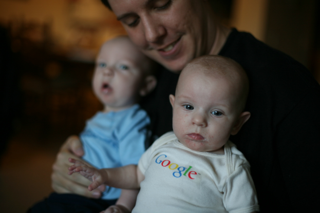 Eine Person hält zwei Babys, mit lesbarem Text auf dem Kleid des vorderen Babys und einem unscharfen Hintergrund.