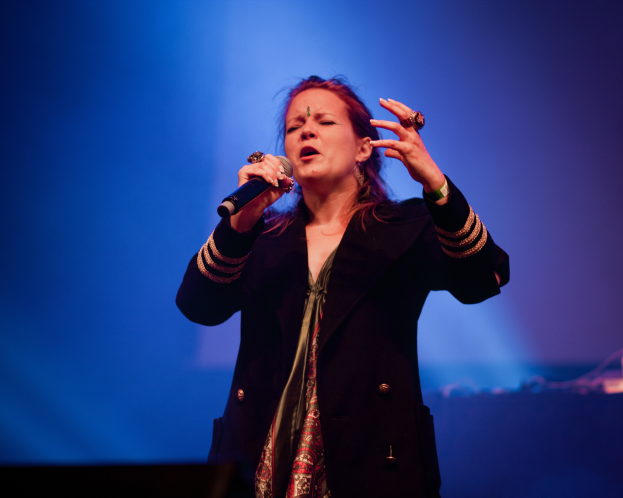 Eine Frau in einem schwarzen Kleid singt auf der Bühne in ein Mikrofon, während sie von blauem Licht beleuchtet wird und ihre Augen geschlossen hat.