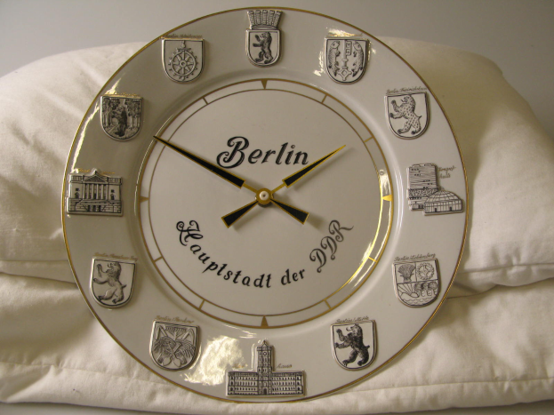 Eine Uhr mit dem Wort 'Berlin' darauf liegt auf einem weißen Kissen an einer Wand im Hintergrund.