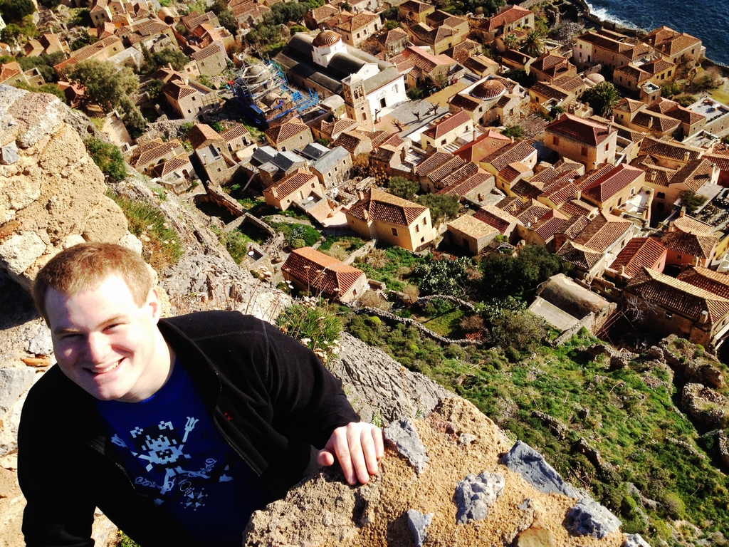 Ein Mann in einer schwarzen Jacke und einem blauen T-Shirt steht links auf dem Bild und lächelt, mit Häusern und Bäumen im Hintergrund.