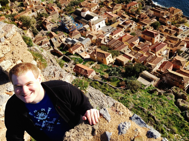Ein Mann in einer schwarzen Jacke und einem blauen T-Shirt steht links auf dem Bild und lächelt, mit Häusern und Bäumen im Hintergrund.