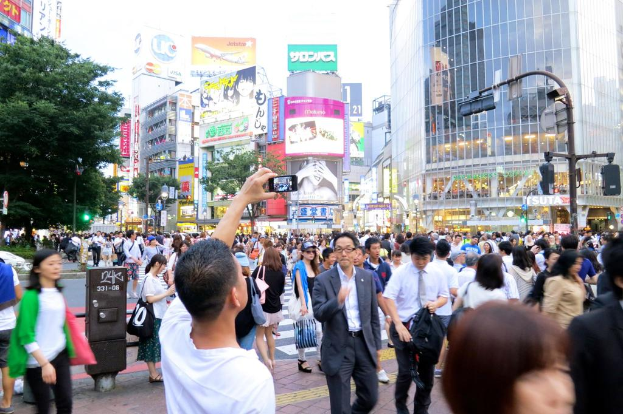 Ein Mann mit einem Mobiltelefon geht auf einem Weg mit anderen Menschen, B√§umen, Pf√∂len, Verkehrszeichen, Geb√§uden und Schildern mit Text im Hintergrund.