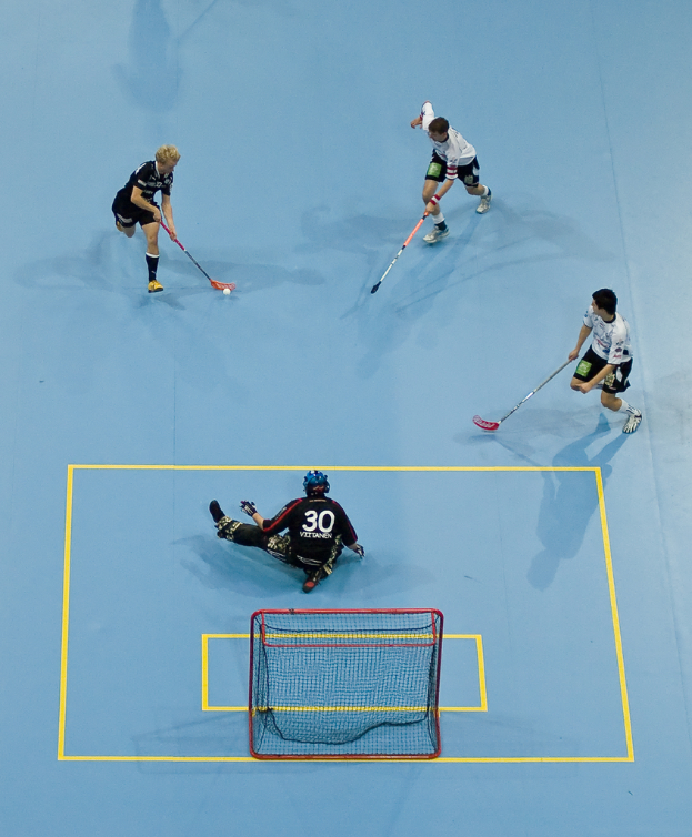 Drei Personen in Sportkleidung spielen Hockey, wobei eine Person rechts den Ball mit einem Hockeystock schlägt und eine Person hinter einem Tor auf dem Boden sitzt.