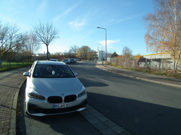 Ein BMW 2er Active Tourer 218d xDrive M Sport fährt auf einer Straße, die von Bäumen, Pfählen, Laternen, Gebäuden und einem Zaun umgeben ist, mit einem klaren blauen Himmel im Hintergrund.