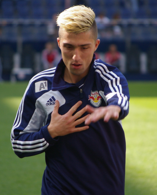 Ein junger Mann in einem blauen T-Shirt steht auf einem Füsballfeld mit ausgestreckten Armen und entschlossener Miene, mit einem unscharfen Hintergrund.