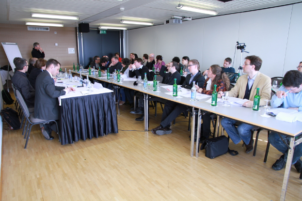 Ein Büroraum mit Mitarbeitern an Tischen, Rucksäcken auf dem Boden, einem Mann im Hintergrund und verschiedenen Gegenständen wie Flaschen, Papieren, einer Kamera, Lautsprecher, Schildern, Projektor und Lampen.